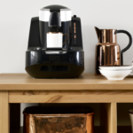 Uma mesa da cor madeira, com um banco da cor cobre embaixo. Em cima da mesa tem uma cafeteira preta grande, alguns potes brancos e um bule de café na cor cobre.